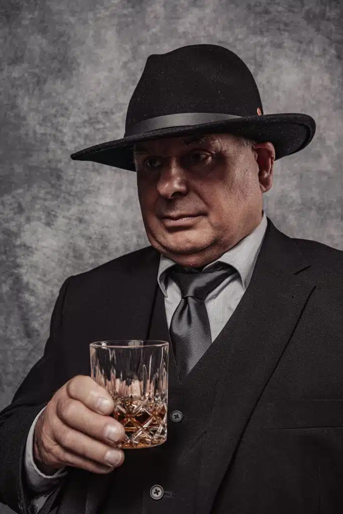 Beeindruckendes Vorher-Nachher-Ergebnis: Professionelles Charakter-Porträt eines Mannes im 1920er-Jahre-Stil mit Hut und Whiskeyglas, fotografiert im Studio in Hamburg-Niendorf.